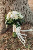 un' nozze mazzo di bianca Rose e verde, decorato con un' seta nastro, sta su un' albero sfondo. nozze particolari. della sposa mazzo foto