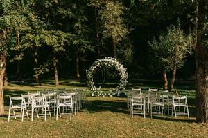 nozze cerimonia nel il foresta. il strada per il il giro arco con fogliame, verdi, verde, e fiori. rustico arredamento. di legno sedie nel il Giardino dietro la casa banchetto la zona. posti a sedere per ospiti. foto