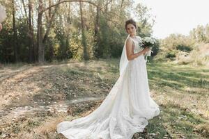 nozze foto nel natura. il sposa è in piedi nel il foresta. il sposa nel un' bellissimo vestito con un' lungo treno, Tenere sua mazzo di bianca Rose, sorridente Cordiali saluti a il telecamera. ritratto