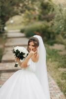 ritratto di il sposa nel natura. un' brunetta sposa nel un' bianca lungo vestire, Tenere un' mazzo di bianca Rose, pose, guardare in il lente, sorridente Cordiali saluti. su il sfondo di il albero. Riccio capelli. foto