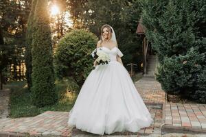 ritratto di il sposa nel natura. un' brunetta sposa nel un' voluminoso vestito e velo, in piedi, in posa con un' mazzo contro il sfondo di un' foresta e un' di legno capanna. un' bellissimo raggio di il sole. foto