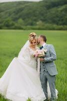 il Novelli sposi In piedi nel un' verde campo e bacio. il sposa è Tenere un' mazzo. nozze nel natura. ritratto di il sposa e sposo. elegante sposo foto