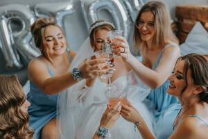 giovane damigelle d'onore nel blu seta vestiti bevanda Champagne nel il della sposa camera. bellissimo donne festeggiare addio al nubilato festa seduta su letto e con Champagne. foto