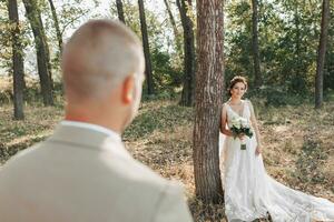 nozze foto nel natura. il sposa è in piedi vicino un' albero nel un' bellissimo vestire, Tenere un' mazzo di bianca Rose, guardare a il sposo. incontro nel il foresta.