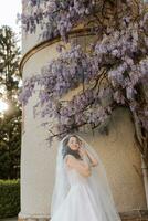 contento giovane donna con lungo Riccio capelli nel un' bianca vestito sotto un' velo vicino un' giardino nel bellissimo fiori. bellissimo ragazza nel il parco foto