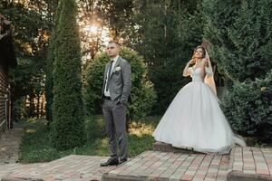 ritratto di il sposa e sposo nel natura. il sposo nel un' grigio completo da uomo è nel il primo piano, il sposa è a piedi dietro a lui, sorridente nel un' bianca voluminoso vestire, Tenere un' mazzo. elegante sposo foto