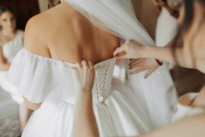 il della sposa amici allacciare il bianca voluminoso vestito di il sposa. posteriore Visualizza. Aperto le spalle. bellissimo mani. lungo velo. mattina di il sposa. foto