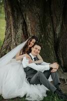 un' bellissimo sposa con lungo Riccio capelli nel un' elegante vestito abbracci il sposo, sorrisi, guardare in il lente sotto un' grande albero. ritratto di il sposa e sposo. primavera nozze. naturale trucco foto