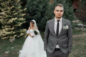 ritratto di il sposa e sposo nel natura. il sposo è nel il primo piano, il sposa è in piedi dietro a lui nel un' bianca voluminoso vestire, Tenere un' mazzo e guardare fuori uso. elegante sposo foto