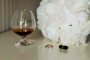 Accessori per il quello dello sposo nozze. un' calice con un' bere, un' bianca mazzo per il sposa, oro anelli, gemelli su un' grigio tavolo. Uomini moda foto