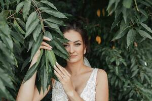 nozze foto su un' verde sfondo. il sposa nel un' bianca vestito sta vicino verde cespugli, detiene un' foglia vicino sua viso e sembra in il lente. bellissimo trucco. ritratto di il sposa