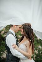 contento giovane coppia. nozze ritratto. il sposa e sposo teneramente bacio sotto un' velo contro il sfondo di un' fioritura cespuglio. nozze mazzo. primavera nozze foto