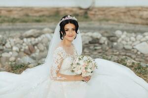 ritratto di il sposa nel natura. un' brunetta sposa con un' corona su sua testa, nel un' bianca voluminoso vestito si siede, pose, Tenere un' mazzo nel sua mani. bellissimo capelli e trucco. nozze tiro foto