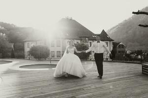 il sposo nel un' nero completo da uomo e il bionda sposa siamo a piedi su il sfondo di il montagna e edifici, vicino il piscina. lungo vestito nel il aria. romantico foto nel natura. bellissimo capelli