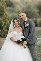 ritratto di il sposa e sposo nel natura. ritagliata foto. il sposa e sposo posa, abbracciare e sorridente, contro il sfondo di conifere. il sposa nel un' lungo vestito con un' mazzo di Rose. foto