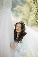 un' bellissimo sposa con lungo Riccio capelli e un' delicato tiara su sua testa. in piedi Il prossimo per il nozze vestire, lei sorrisi Cordiali saluti. ritratto di un' ragazza guardare in il lente. naturale trucco foto