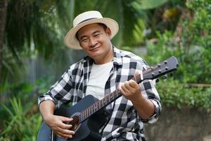 bello asiatico uomo è giocando acustico chitarra nel il parco, all'aperto natura sfondo. concetto, amore musica, passatempo, ricreazione attività. rilassare volta. musica può sviluppare mentale Salute. foto