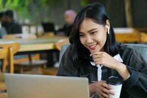 eccitato asiatico donna bevanda caffè e utilizzando il computer portatile per avere in linea incontro nel bar, digitale nomade foto