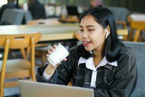 allegro asiatico donna godere bevanda caffè e utilizzando il computer portatile per avere in linea incontro nel bar foto