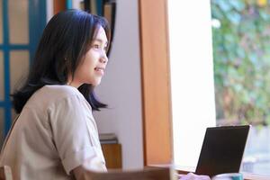 contento asiatico donna Lavorando su il computer portatile seduta vicino finestra di caffè negozio indossare casuale Abiti, femmina progettista avere in linea incontro progetto nel bar foto