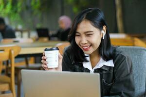 allegro asiatico donna Tenere un' tazza di caffè e utilizzando il computer portatile per avere in linea incontro nel bar foto