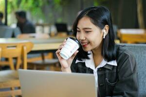 eccitato asiatico donna bevanda caffè e utilizzando il computer portatile per avere in linea incontro nel bar, digitale nomade foto