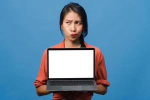 la giovane donna asiatica mostra lo schermo del laptop vuoto con un'espressione positiva, sorride ampiamente, vestita con abiti casual sentendo felicità isolata su sfondo blu. computer con schermo bianco in mano femminile. foto