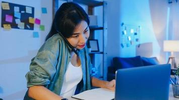 donne asiatiche freelance che utilizzano laptop parlano al telefono imprenditore impegnato che lavora a distanza nel soggiorno. lavoro da sovraccarico domestico di notte, lavoro a distanza, distanza sociale, quarantena per coronavirus.