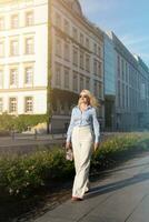 un' elegante, positivo donna nel i pantaloni e un' camicia a piedi giù il strada vicino un' europeo costruzione. un' caldo estate giorno foto