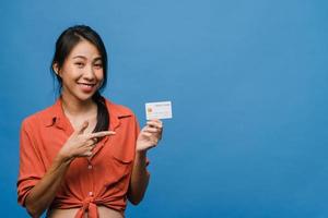 la giovane donna asiatica mostra la carta di credito con l'espressione positiva, sorride ampiamente, vestita con abiti casual sentendo felicità e stando isolata su sfondo blu. concetto di espressione facciale. foto