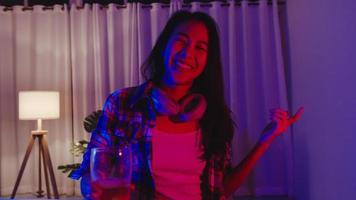 giovane signora asiatica che beve birra divertendosi momento felice discoteca neon night party evento celebrazione online tramite videochiamata nel soggiorno di casa. distanziamento sociale, quarantena per la prevenzione del coronavirus.