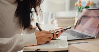 primo piano giovane donna asia freelance focus telefono cellulare chat con i colleghi lavoro finanza scrivendo sul taccuino nel soggiorno di casa. studentessa impara online a casa, lavora da casa concetto. foto