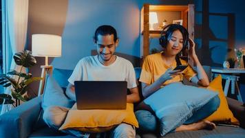 freelance asia coppia uomo e donna in casual hasband lavoro computer portatile di notte godersi felice relax con moglie indossare cuffie ascoltare musica in smartphone cantare danza sul divano a casa, vita coniugale. foto