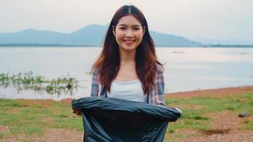 il ritratto di una giovane donna asiatica volontaria aiuta a mantenere la natura pulita guardando la macchina fotografica e sorride con i sacchetti della spazzatura neri sulla spiaggia. concetto sui problemi di inquinamento di conservazione ambientale. foto
