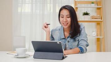 donne d'affari freelance abbigliamento casual utilizzando tablet videoconferenza chiamata di lavoro con il cliente sul posto di lavoro nel soggiorno di casa. felice giovane ragazza asiatica rilassarsi seduto sulla scrivania fare un lavoro in internet. foto