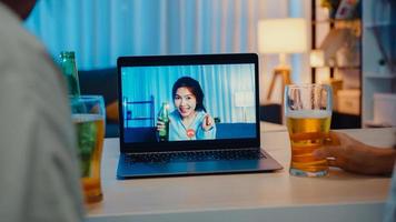 giovane donna asiatica che beve birra divertirsi momento felice festa notturna evento celebrazione online tramite videochiamata nel soggiorno di casa di notte. distanziamento sociale, quarantena per la prevenzione del coronavirus.