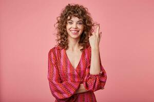 giovane, Riccio donna sta al di sopra di il rosa sfondo, sorridente e torsione capelli su dito, indossare romantico a strisce vestito con un' modello foto