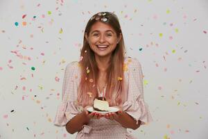 contento giovane bellissimo lungo dai capelli femmina mostrando sua piacevole emozioni mentre festeggiare sua compleanno, sorridente positivamente con torta nel sollevato mani, in posa al di sopra di bianca sfondo foto