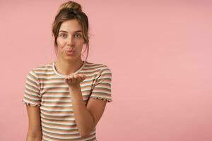 studio foto di giovane bella Marrone dai capelli signora con ciambella acconciatura raccolta sua palma e pieghevole labbra nel aria bacio mentre guardare positivamente a telecamera, isolato al di sopra di rosa sfondo