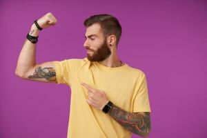 forte attraente giovane tatuato maschio con barba e di moda taglio di capelli guardare con orgoglio a il suo mano e dimostrando il suo bicipite, in piedi al di sopra di viola sfondo nel casuale maglietta foto