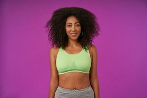 studio tiro di sportivo giovane attraente buio spellato femmina con Riccio capelli indossare leggero verde superiore, in piedi al di sopra di viola sfondo con mani fuori uso, guardare a telecamera con sincero Sorridi foto