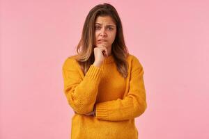 confuso giovane occhi verdi brunetta femmina conservazione mano su sua mento e raccolta sopracciglio mentre guardare a telecamera, pungente labbro inferiore mentre in posa al di sopra di rosa sfondo foto