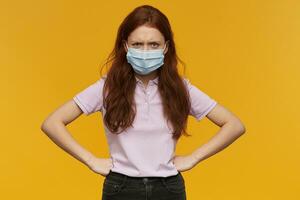 pazzo irritata giovane donna indossare medico protettivo maschera mantiene mani su vita e guardare a telecamera al di sopra di giallo sfondo foto