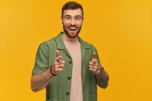 allegro attraente barbuto giovane uomo con tatuaggio sorridente e puntamento su voi a telecamera di Due dita al di sopra di giallo sfondo foto
