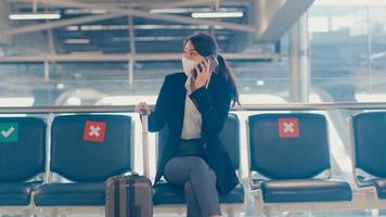 smart asian business girl indossare tuta seduta con valigia in panchina chiamata smart phone parlare con il partner in attesa del volo in aeroporto. pendolare di viaggio d'affari nella pandemia di covid, concetto di viaggio d'affari. foto