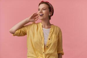 Immagine di europeo affascinante donna 20s con naturale trucco Marrone con gli occhi indossare estate elegante attrezzatura giallo camicia e rosso bandana in posa contro rosa parete emozione gioia viso guardare a parte dita toccare orecchio foto