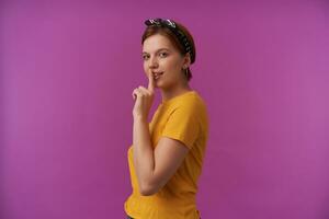 Immagine europeo Marrone dai capelli affascinante donna 20s con naturale trucco indossare elegante estate giallo maglietta con braccia al di sopra di viso dito toccare labbra emozione abbastanza silenzio segreto in posa contro viola parete foto