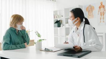 la giovane dottoressa asiatica indossa una maschera protettiva usando gli appunti sta offrendo grandi discorsi di notizie discutere risultati o sintomi con la ragazza paziente nell'ufficio dell'ospedale. stile di vita nuovo normale dopo il virus corona. foto