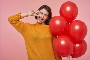 interno ritratto di giovane positivo femmina indossa rosso luminosa maglione sorridente e mostrando v gesto. isolato al di sopra di rosa sfondo foto