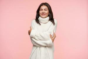 piacevole guardare giovane bella Marrone dai capelli donna conservazione sua occhi chiuso mentre abbracciare lei stessa con sollevato mani e sorridente allegramente, in posa al di sopra di rosa sfondo foto